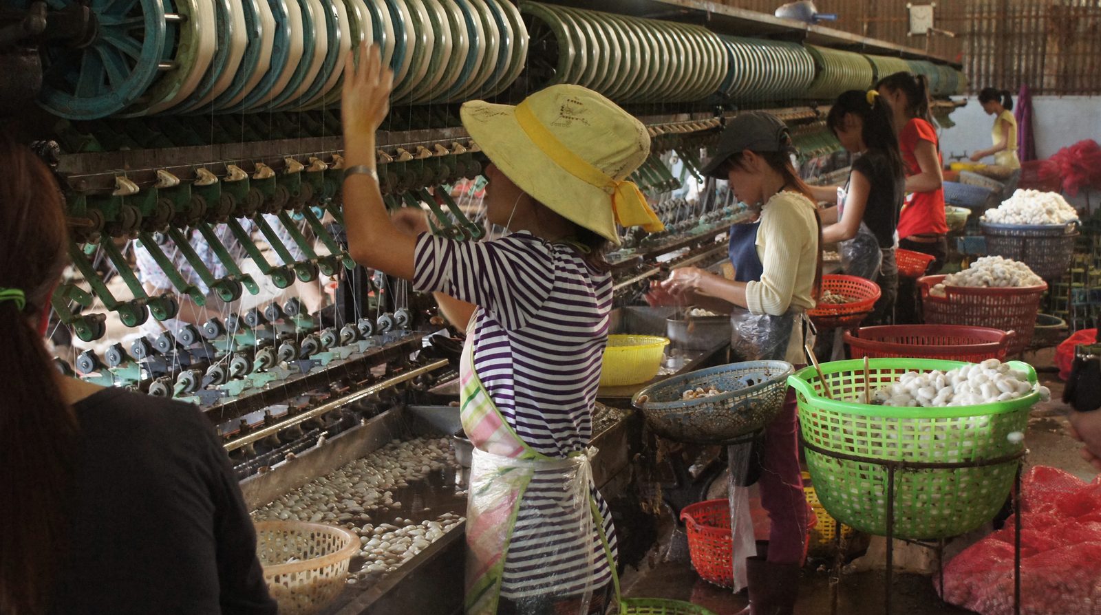 Công nhân đứng máy kéo sợi tơ trong một xưởng dệt lụa ở Việt Nam, nhiều sọt kén tằm xếp trước máy