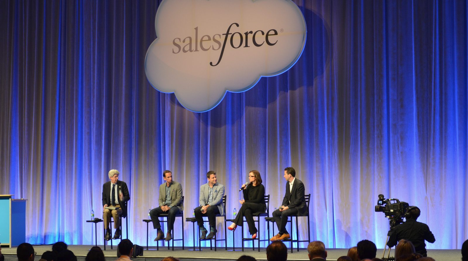 Five speakers on tall chairs on a stage with the Salesforce cloud logo and blue curtains, with the audience and a camera operator in front